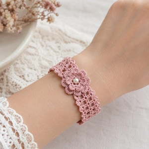 May include: A dusty pink crochet bracelet featuring a central flower motif with a small white pearl at its core. The band is constructed from a delicate, open-work lattice pattern, creating a soft and intricate texture that wraps around the wrist.