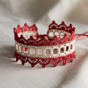 Pode incluir: Uma pulseira de crochê vermelha e creme feita à mão. A pulseira apresenta uma borda ondulada e um design vazado. Os detalhes intrincados do trabalho de crochê são visíveis, mostrando o artesanato. A pulseira é exibida em uma superfície branca.