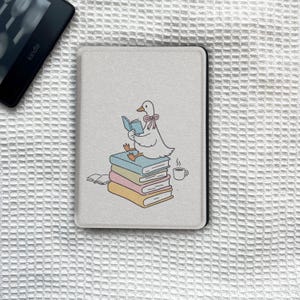 May include: A light-coloured Kindle cover with a cartoon duck reading a book on a stack of colourful books. A small cup of coffee is next to the books. The cover is rectangular with rounded corners.