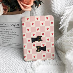 Peut inclure: Un tapis rectangulaire à carreaux roses et blancs avec des cœurs rouges et deux illustrations de chats noirs. Le tapis est sur une surface blanche, avec un sac en papier et des fleurs en arrière-plan.