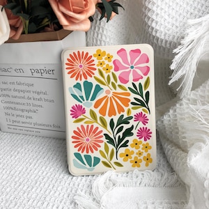 Op de afbeelding: Een rechthoekige telefoonhoes met een bloemenpatroon. De hoes heeft een crèmekleurige achtergrond met kleurrijke bloemen in tinten oranje, roze, geel en blauwgroen, en groene bladeren. De hoes is waarschijnlijk gemaakt van een duurzaam materiaal.