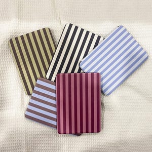 Op de afbeelding: Vijf rechthoekige notitieboekjes met verticale strepen in verschillende kleuren: bruin en beige, zwart en wit, blauw en wit, paars en blauw en bordeauxrood en roze. De notitieboekjes liggen op een wit, gestructureerd oppervlak.