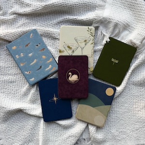 Può includere: Sei custodie per e-reader con vari design: una blu con cani che nuotano, una crema con bicchieri da martini, una verde scuro con una libellula, una viola scuro con un cigno, una blu navy con una stella e una con un paesaggio.