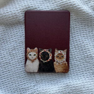 May include: A rectangular, burgundy-colored case featuring a whimsical illustration of three cats. Each cat wears a bonnet with a floral pattern. The cats are white, black, and orange tabby.