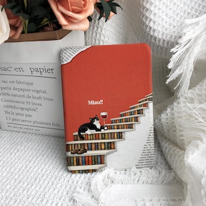 May include: A red notebook with a black and white cat illustration, a glass of wine, and the word "Miau!!". The cat is lying on a staircase made of bookshelves. The notebook has a paper corner.