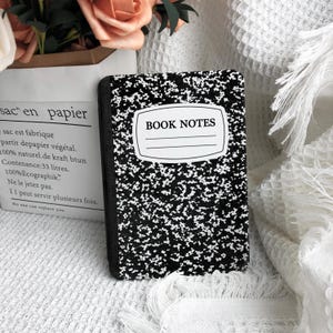 May include: A black and white composition notebook with the words "BOOK NOTES" on the cover. The notebook has a speckled design and a white label. The notebook is open and ready to be written in.