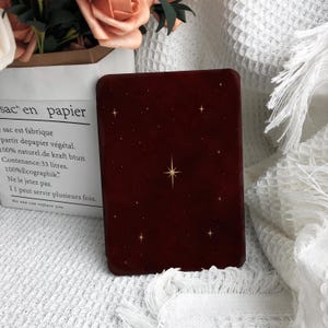 May include: A deep burgundy e-reader cover featuring a gold starburst pattern across the front surface. The cover has a smooth, matte finish and is positioned upright. The design includes various small gold stars scattered across the dark red background, with one larger, central starburst as the focal point of the accessory.