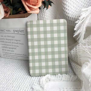 Puede incluir: Una funda rectangular para cuaderno o tableta con un patrón de cuadros vichy verde claro y blanco. La funda está sobre una superficie blanca texturizada, con una bolsa de papel y flores en el fondo.