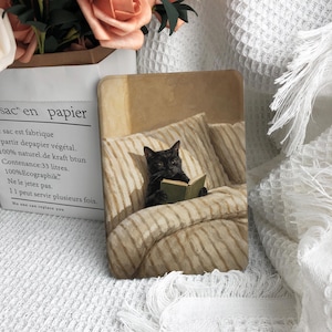 May include: A rectangular print featuring a black cat sitting upright in a bed with beige and white striped pillows and a matching blanket. The cat is holding an open green book with both paws. The scene is rendered in a soft, painterly style with warm, muted tones, capturing the cat in a relaxed, focused posture while reading in the cozy, textured bedding.