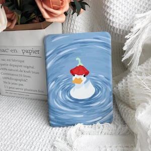 May include: A rectangular, blue-toned case featuring a whimsical illustration of a white duck wearing a red hat, floating in water. The duck has an orange beak and is surrounded by concentric blue circles. The case is a unique accessory.
