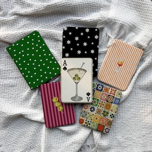 Può includere: Una collezione di sottobicchieri decorativi con vari disegni. I sottobicchieri includono uno verde con cuori bianchi, uno nero con fiori bianchi e uno arancione a righe con un bicchiere da cocktail. Altri sottobicchieri presentano un martini e un motivo floreale.