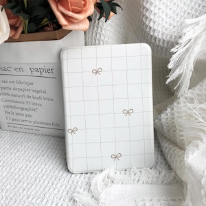May include: A white rectangular case with a grid pattern and small bow accents. The case is likely for a phone or tablet, and the design is simple and elegant. The background includes a paper bag with text and flowers.