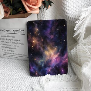 May include: A rectangular e-reader cover featuring a vibrant galaxy print with deep purple, blue, and gold nebula clouds scattered with bright white stars against a dark background. The cover has rounded corners and a smooth, flat surface.