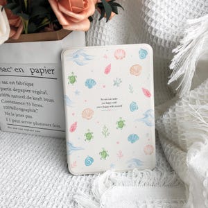 Könnte beinhalten: Eine weiße E-Reader-Hülle mit einem wiederkehrenden Muster aus aquarellartigen Meeresschildkröten, verschiedenen Muscheln, Korallen und blauen Wellen. In der Mitte befindet sich der aufgedruckte Text: No one can make you happy until you're happy with yourself.