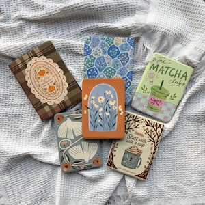 Può includere: Sei custodie rettangolari per e-reader con vari design: una custodia a quadri marroni con etichetta a zucca, un motivo floreale blu, una custodia verde con la scritta The Matcha Club, una grigia con uccelli bianchi, una arancione con margherite e una color crema.