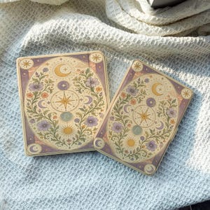 Puede incluir: Dos estuches rectangulares con un diseño celestial que presenta motivos centrales de estrella, sol y luna rodeados de flores moradas y naranjas. El fondo es de color crema con bordes morados, decorado con pequeñas estrellas y lunas.