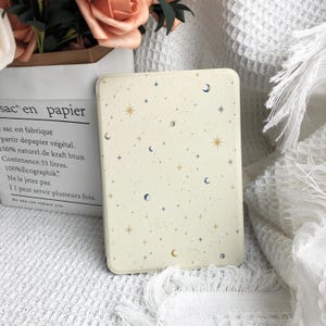 May include: A cream-colored e-reader case featuring a celestial pattern of small gold and blue stars, crescent moons, and tiny dots. The rectangular cover has rounded corners and a smooth, matte finish, displaying a consistent cosmic print.