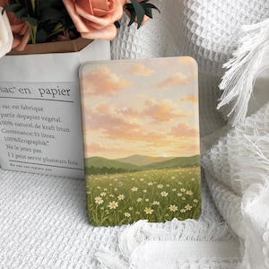 May include: A rectangular e-reader case featuring a printed landscape illustration of a green meadow filled with white daisy flowers, rolling hills in the distance, and a soft sunset sky with pink and orange clouds. The case has a smooth finish.