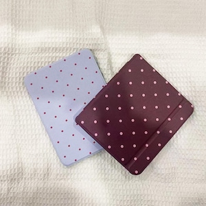 Puede incluir: Dos fundas cuadradas con esquinas redondeadas. Una es azul claro con pequeños lunares rosa oscuro. La otra es de color burdeos intenso con lunares rosa claro. Ambas están sobre una superficie blanca texturizada.