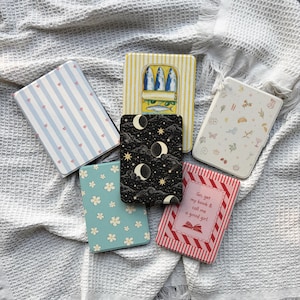 May include: A collection of decorative, rectangular cases with various designs. The cases feature patterns such as blue stripes with hearts, a sardine can, a night sky with moons, floral patterns, and text that reads "Go, get my book & call me a good girl."