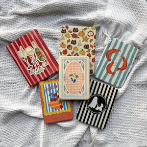 May include: Five rectangular e-reader cases featuring various designs: red stripes with champagne glasses and the text "drink & be merry," a cream background with autumn motifs, blue stripes with shrimp, a cherry bowl with "Summer Fruit Club" text, and black stripes with a ghost and dog.