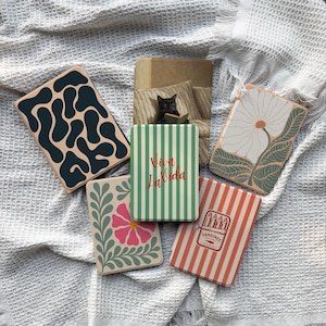 May include: A collection of decorative coasters with various designs. One coaster features a dark teal abstract pattern on a beige background. Another has a floral design with a pink flower and green leaves. Other coasters have text and striped patterns.
