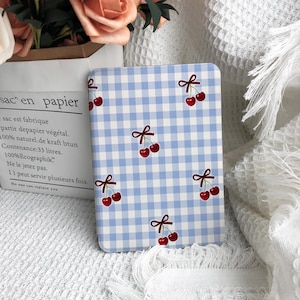 May include: A rectangular, light blue and white checkered pattern case with a repeating pattern of red cherries and small bows. The case is on a white surface with a paper bag and flowers in the background.