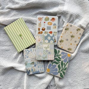 May include: Six rectangular e-reader or tablet cases featuring various designs: one with green stripes and a palm tree, one with breakfast icons, one with palm trees, one with a floral patchwork, one with a coastal scene labeled East Coast, and one with large blue flowers.