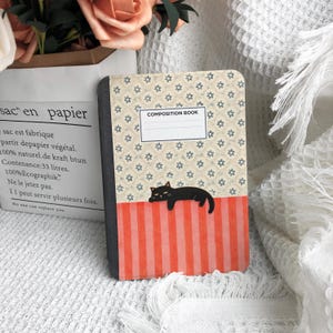 May include: A composition book with a patterned cover featuring a black cat and the words "COMPOSITION BOOK". The top half of the cover has a floral design, while the bottom half has orange and white stripes.