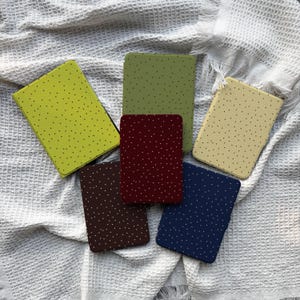 Op de afbeelding: Een verzameling van zes rechthoekige hoesjes in verschillende kleuren, waaronder groen, bordeauxrood en marineblauw. Elk hoesje is bedekt met een patroon van kleine, gelijkmatig verdeelde witte stippen. De hoesjes liggen op een witte, getextureerde ondergrond.