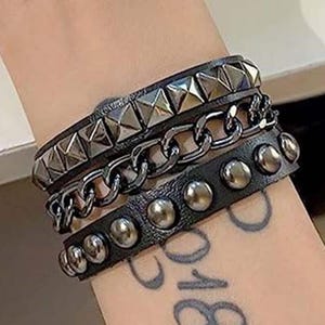 Puede incluir: Un conjunto de pulseras de cuero negro con detalles metálicos plateados. La pulsera superior presenta tachuelas piramidales, la del medio un diseño de cadena y la inferior tachuelas redondas. Las pulseras están apiladas.