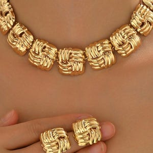 Puede incluir: Un conjunto de joyas doradas que incluye un collar y pendientes a juego. El collar y los pendientes tienen un diseño cuadrado tejido. Las joyas se exhiben sobre la piel de una persona, resaltando el color y la textura de las piezas.