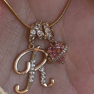May include: Gold-coloured necklace with a pendant featuring the letter "R" and a pink heart. The letter and heart are adorned with clear crystals. The necklace chain is a thin, textured gold. The pendant is suspended from a delicate chain.