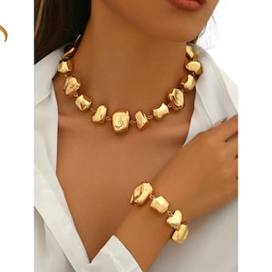 Puede incluir: Un conjunto de collar y pulsera en tono dorado. La joyería presenta formas geométricas irregulares y gruesas. El collar está alrededor del cuello y la pulsera en la muñeca. La imagen también incluye el texto "Lakech Jewelry" en dorado.