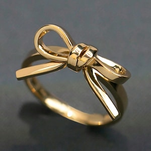 May include: A gold-colored ring with a bow design. The ring features a detailed bow shape at the top, with the band curving around to form the ring. The ring is made of a shiny, reflective metal.