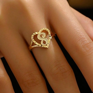 May include: A gold-colored ring featuring a heart-shaped design with an openwork pattern. The ring has a capital letter "S" in the center of the heart. The ring is worn on a finger.