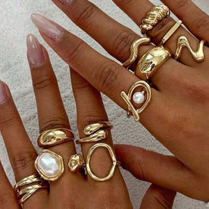 Könnte beinhalten: Nahaufnahme von Händen, die mit verschiedenen goldenen Ringen geschmückt sind. Die Ringe weisen unterschiedliche Designs auf, darunter ein Perlenakzent, verdrehte Bänder und glatte, abgerundete Formen. Die Nägel sind mit hellem rosa Nagellack lackiert.