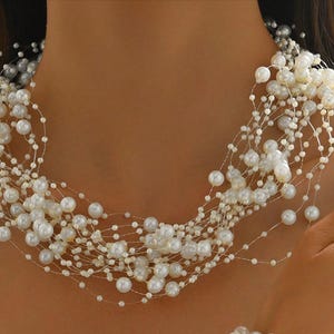 May include: A multi-strand pearl necklace featuring off-white pearls of different sizes. The necklace is draped around the neck and shoulders, creating a cascading effect. The pearls are strung on delicate, nearly invisible strands.