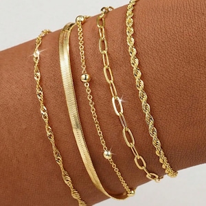 May include: A collection of five gold bracelets, each with a unique design, adorns a wrist. The bracelets include a twisted rope chain, a flat herringbone chain, a delicate chain with small beads, a chain with oval links, and a thicker rope chain.