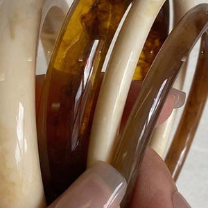 May include: A collection of chunky bangles in shades of cream, amber, and brown. The bracelets are made of a glossy material and are stacked together, showcasing their varying colors and textures. The bangles are held in a hand.