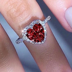 Puede incluir: Un anillo de plata con una piedra preciosa roja oscura en forma de corazón. El corazón está rodeado por un halo de piedras más pequeñas y transparentes. El anillo está en un dedo, mostrando los detalles brillantes de la joya.