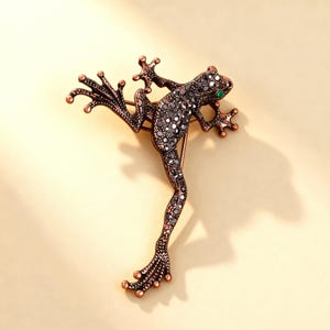 以下が含まれることがあります： 銅色の金属製カエルのブローチ。質感のある仕上げが施され、体には多数の小さな暗い反射石が埋め込まれています。カエルには明るい緑色の石の目が一つあり、手足は広がり、水かきのある足が詳細に表現されています。