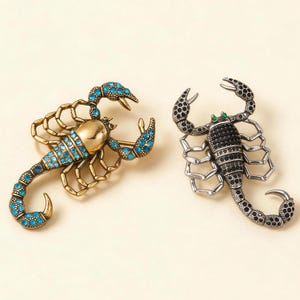 Puede incluir: Dos broches con forma de escorpión. El broche de la izquierda es de tono dorado con pedrería azul brillante en la cola y las pinzas. El broche de la derecha es de tono plateado con pedrería negra en el cuerpo y la cola, con dos ojos verdes.