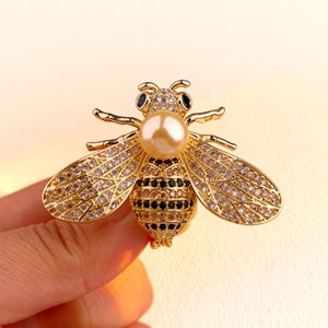 Puede incluir: Broche de abeja dorada con cuerpo de perla y alas con incrustaciones de cristal. El broche presenta detalles negros en el abdomen y los ojos. Esta joya es un accesorio decorativo.