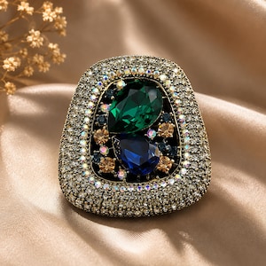 May include: A brooch featuring a large pear-shaped emerald green crystal and a pear-shaped sapphire blue crystal at the center. The piece is surrounded by a wide border of small clear crystals and accented with smaller amber and dark blue stones.
