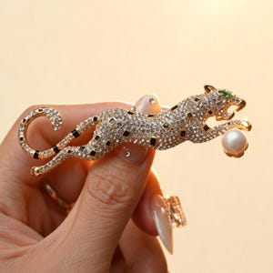 May include: A gold-toned leopard brooch encrusted with clear crystals and black enamel accents. The leopard is depicted in a leaping pose, holding a single white spherical pearl in its front paws, and features a small green crystal for its eye.
