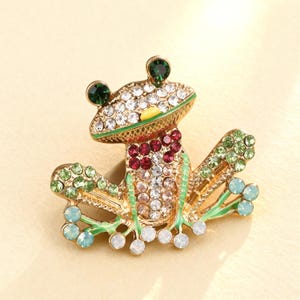 Könnte beinhalten: Eine goldfarbene Froschbrosche, besetzt mit klaren, grünen, roten und blauen Kristallen. Der Frosch hat grüne Augen, eine Brust aus roten und klaren Kristallen und hellblaue und weiße Kristallakzente an den Füßen.