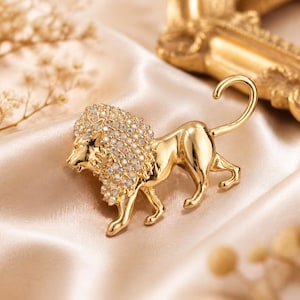 May include: A gold-toned lion brooch featuring a mane encrusted with iridescent, multi-faceted crystals. The lion is depicted in a standing profile position with a curved tail, smooth polished metal body, and four legs visible in a walking stance.