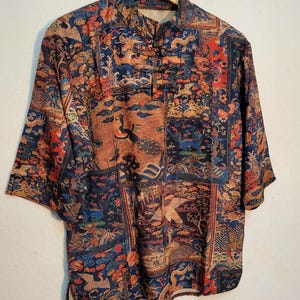 May include: A short-sleeved shirt with a mandarin collar and a button-down front. The shirt features a patchwork design with a variety of patterns in shades of blue, brown, orange, and red. The shirt is hanging on a wooden hanger.
