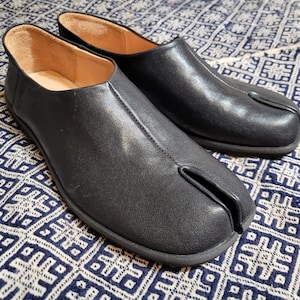 May include: A pair of black leather split-toe shoes, also known as Tabi shoes, with a low profile and a slip-on design. The shoes have a visible seam and a rubber sole. The interior is a light tan color. The shoes are displayed on a patterned fabric.
